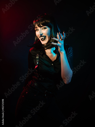a girl with vampire fangs with black hair in a black shirt and harness raised her hand with long nails in a different-colored light