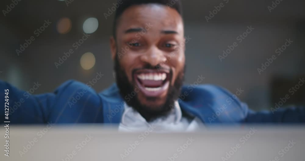 Cheerful businessman celebrating with arms raised sitting at desk in office and working on laptop