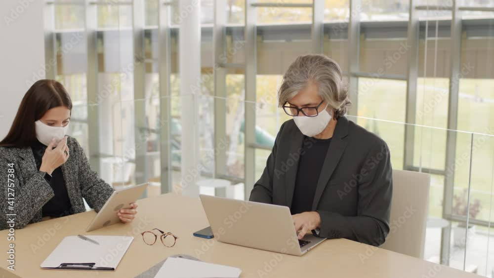 Slow-motion panning medium shot of adult coworkers wearing protective masks sitting at table in modern building using laptop and digital tablet working Slow-motion panning medium shot of adult coworkers wearing protective masks sitting at table in modern building using laptop and digital tablet working