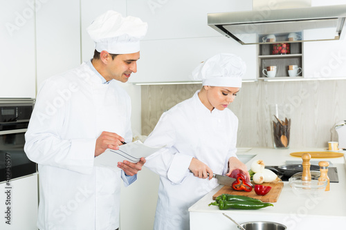 Positive female and male young cooks with paper recipe in uniform working on kitchen