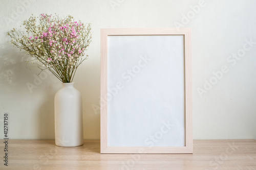 Portrait white picture frame mockup on wooden table. Modern ceramic vase with gypsophila.  White wall background. Scandinavian interior. Vertical. 