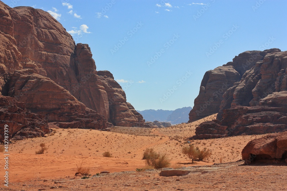 canyon between the relief red mountains, dry lifeless trees grow on the sand, small car rides in the canyon, clear day, wadi rum desert, jordan
