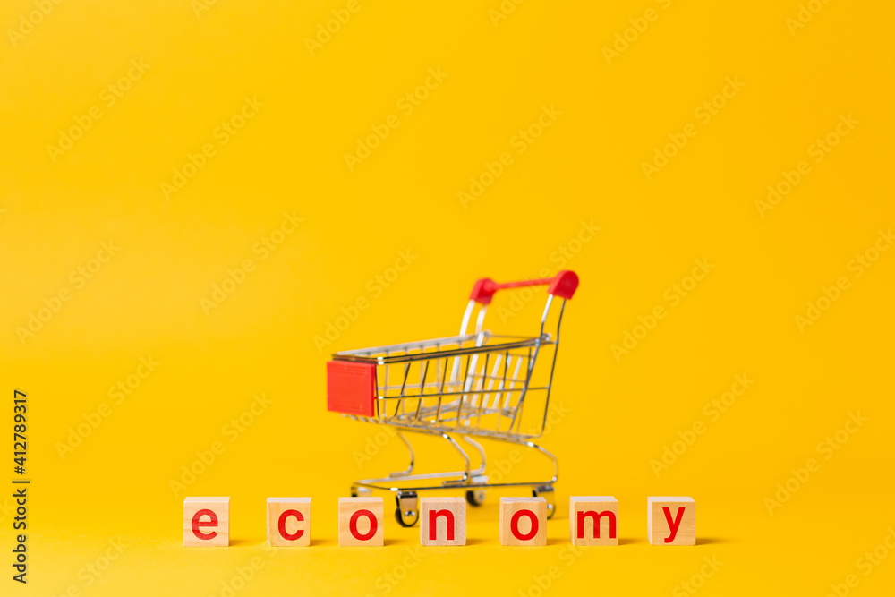 An empty cart and wooden blocks, with a red text economy. Yellow background. Copy space. The concept of economy, crisis and shopping