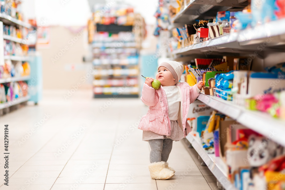 Shopping. A cute baby girl stands at the supermarket shelves with a pear in his hand. The concept of shopping and consumerism