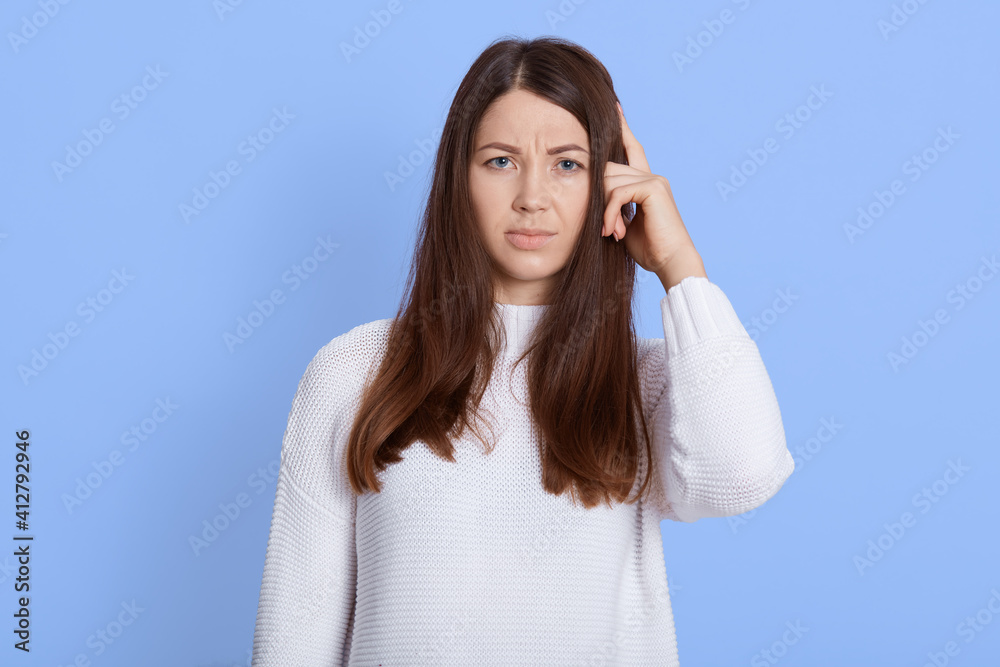 Portrait of forgetful female dark haired female remembers something, keeps finger on temple, remembers to do important task, wears white casual sweater, looks at camera against blue wall. Portrait of forgetful female dark haired female remembers something, keeps finger on temple, remembers to do important task, wears white casual sweater, looks at camera against blue wall.