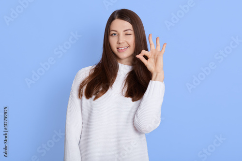 Young woman showing Ok sign isolated over blue background, dark haired lady winking eye and smiles while looking directly at camera, female with pleasant appearance wearing white casual shirt. © sementsova321
