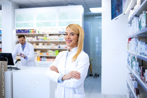 Portrait of professional woman pharmacist proudly standing in pharmacy shop or drugstore. In background shelves with medicines. Healthcare and medicine.