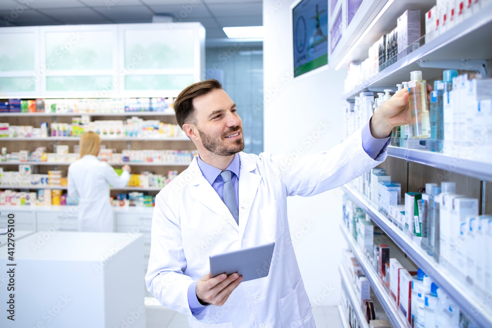 Professional handsome male pharmacist working in pharmacy store or drugstore. Taking medicines of the shelf. Healthcare and apothecary.