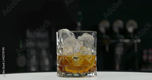 Rocks glass with golden whisky with lemon in bar. Unrecognizable bartender putting ice cubes in alcoholic drink. Preparing whiskey sour cocktail on the rocks. Bokeh effect