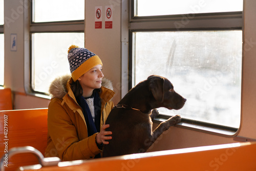 Young happy woman wear winter clothes sitting in local train with her lovely dog, hugging, thinking, looking through the window, traveling together. Love pets. 