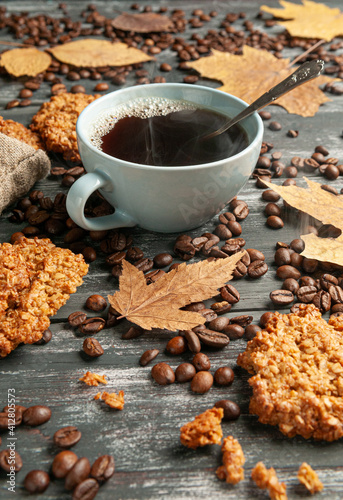 Coffee in a blue cup. Homemade oatmeal cookies are nearby. Decorated with coffee beans and yellow leaves. Dark wooden background.
