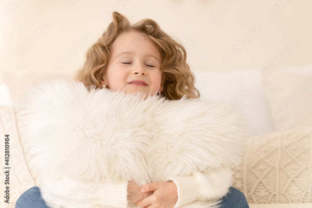 Cute little girl with long blond hair hugging a pillow with close eyes and smiling. Happy child at home in beige interior. closeup