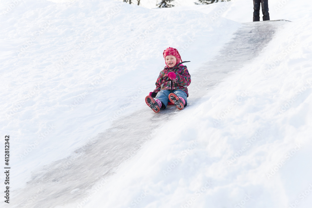 Winter activities outdoors. Cute happy preschool girl wearing a warm clothes is having fun, sliding down a hill on the ice slide