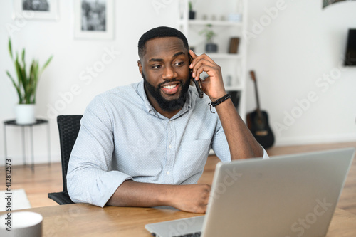 Surprised Afro American young man looking at the screen of the laptop, talking on the phone, printing, enjoing freelance work online. Front view portrait light home office. Business conversation