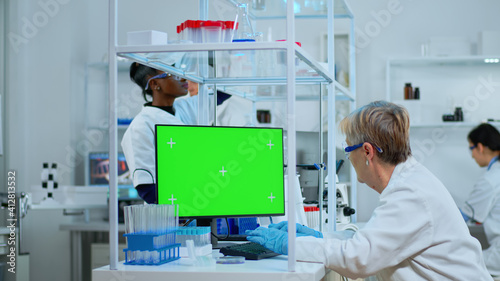 Senior doctor working at computer with green screen in modern equipped lab. Multiethnic team of microbiologists doing vaccine research writing on device with chroma key  isolated  mockup display.