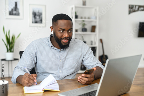Focused happy young african american man student freelancer making notes studying working with laptop, wearing airpods looking at the screen of the laptop, chatting on the phone at home office