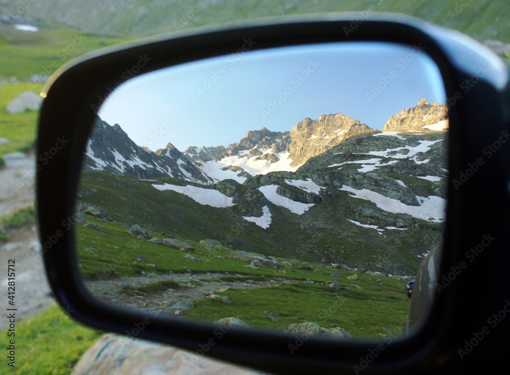 A close up shot of a side mirror which reflects the green grass and the mountain filled with snow on it.