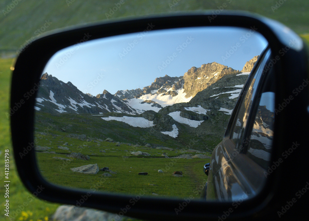 A close up shot of a side mirror which reflects the green grass and the mountain filled with snow on it.