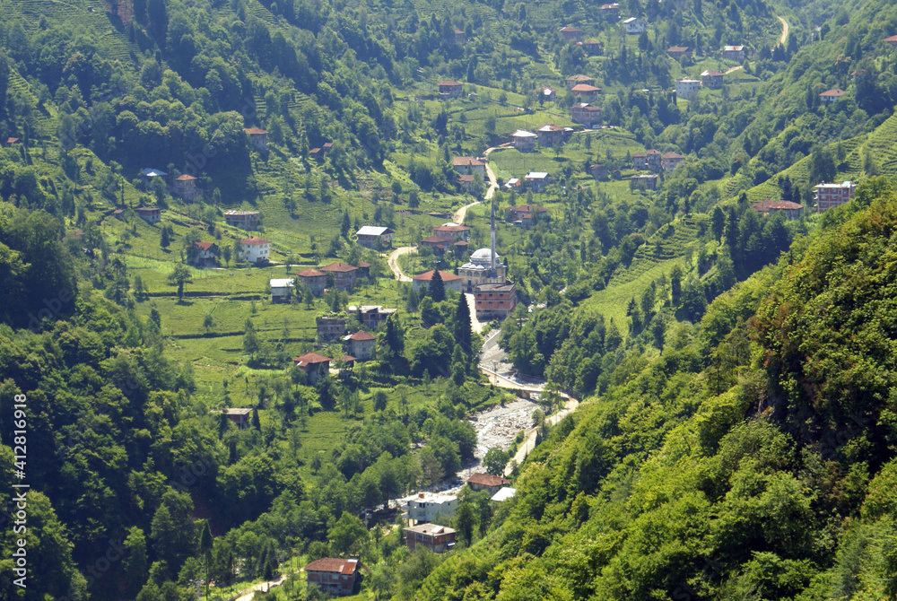 A photo of nature. This is a panoramic shot of valleys filled with green grass, trees, light coloured houses and a mosque near a path that goes up.	