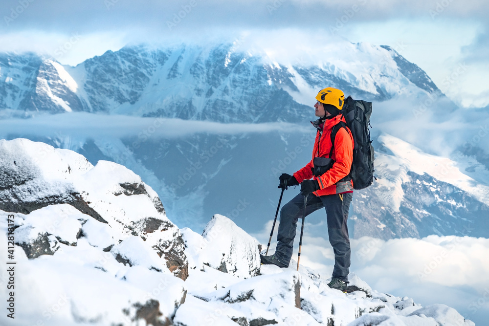 A middle aged man climber with a large backpack on his shoulders walks up a stone slope against the backdrop of beautiful snow capped mountains with glaciers