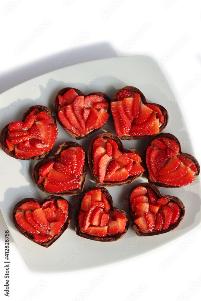 Fototapeta premium Valentine’s day dessert background. Mini heart shaped chocolate tarts with fresh strawberries on a plate isolated on white