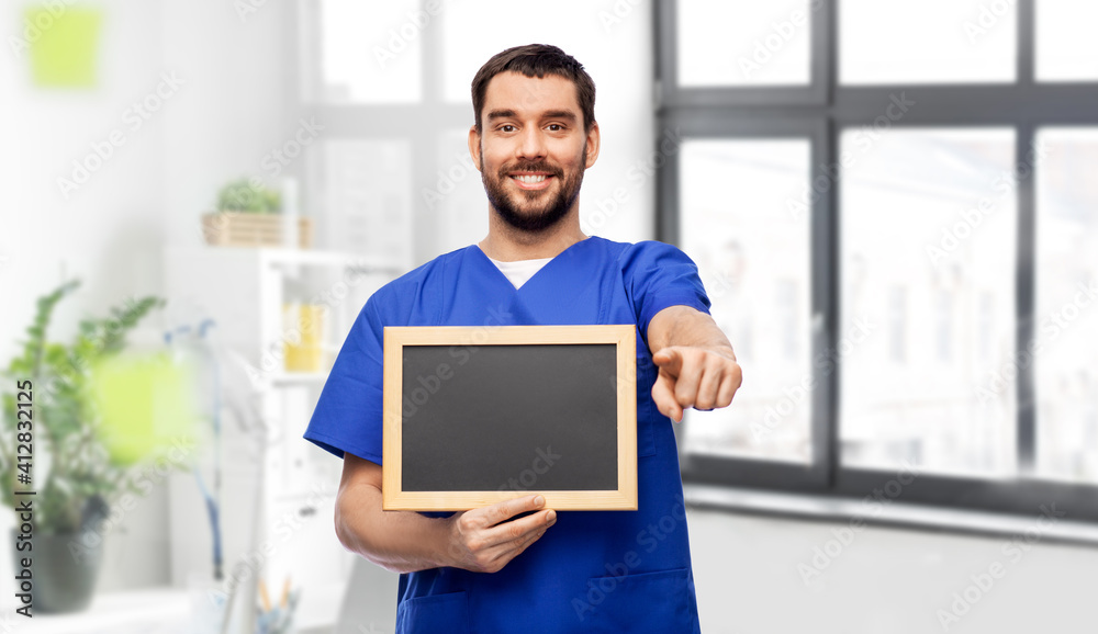 healthcare, profession and medicine concept - happy smiling male doctor or nurse in blue uniform with chalkboard pointing finger to camera over medical office at hospital background