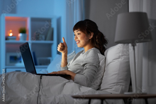 technology, internet, communication and people concept - happy smiling young african asian with laptop computer having video call in bed at home at night and showing thumbs up