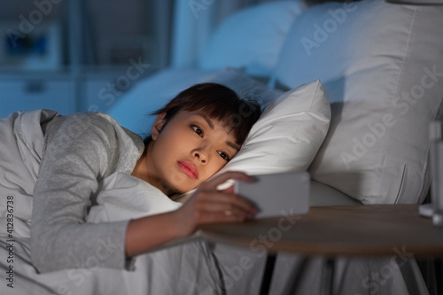 technology, internet, communication and people concept - young asian woman with smartphone lying in bed at home at night