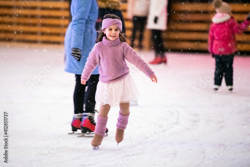 little girl in a pink sweater and skirt is skating on a winter evening on an outdoor ice rink lit with garlands