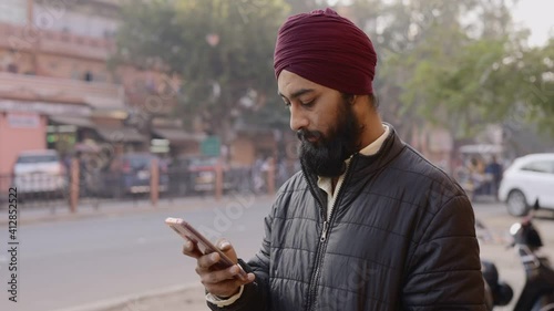 shot of a young Indian office worker man wearing turban on head with long beard using a mobile phone standing by the corner of a street during morning hours