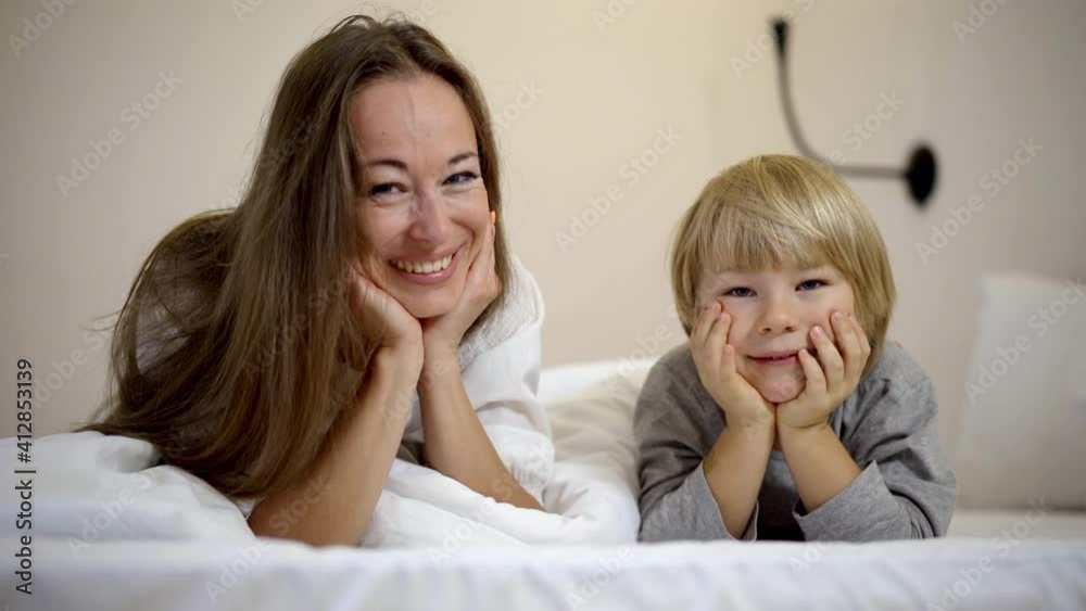 Portrait of happy mother and son looking at camera smiling holding face on hands. Cheerful beautiful Caucasian cute boy with blue eyes posing in bedroom at home in the morning. Portrait of happy mother and son looking at camera smiling holding face on hands. Cheerful beautiful Caucasian cute boy with blue eyes posing in bedroom at home in the morning.