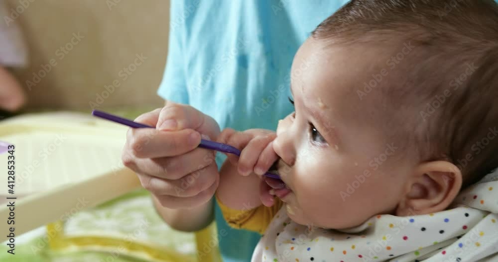 mother holds the baby in her arms and feeds her boy with a spoon. family, food, child, eating and parenthood concept. slow motion mother holds the baby in her arms and feeds her boy with a spoon. family, food, child, eating and parenthood concept. slow motion