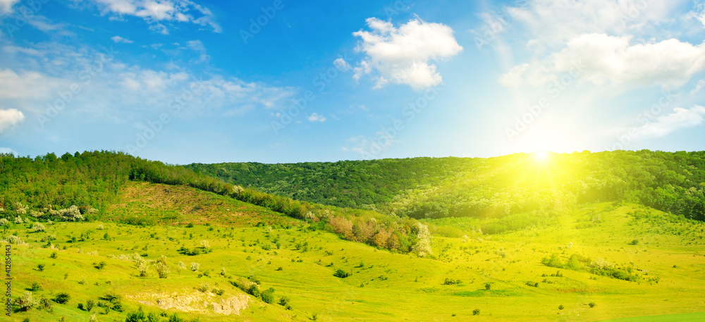 Green grass field on hills and sun on blue sky . Wide photo.