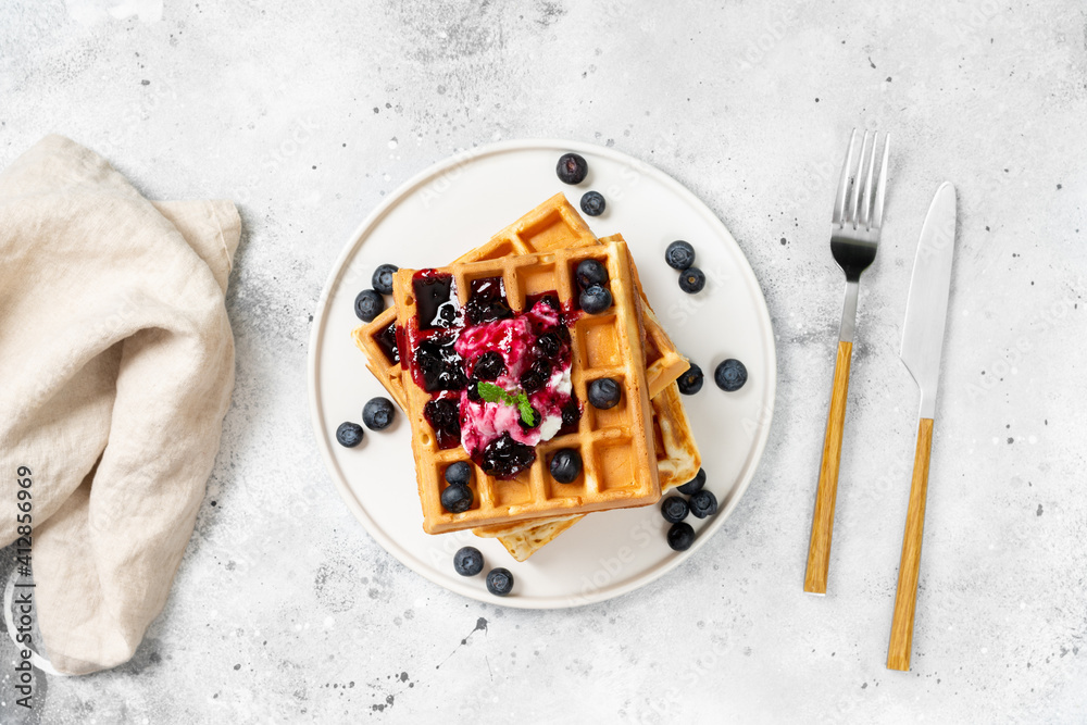 Belgian or Brussels waffles with Greek yogurt and blueberry jam. Homemade waffles in a white plate on the light gray kitchen table. Traditional classic Belgian waffles. Top view