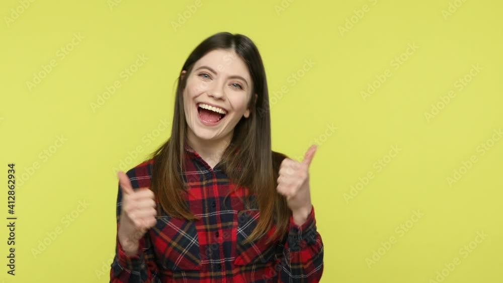 Positive excited brunette woman in checkered shirt showing thumbs up like gesture looking at camera with toothy smile, satisfied with service, rating. Indoor studio shot isolated on yellow background Positive excited brunette woman in checkered shirt showing thumbs up like gesture looking at camera with toothy smile, satisfied with service, rating. Indoor studio shot isolated on yellow background