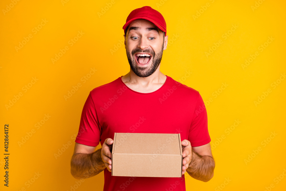 Photo portrait of funny laughing man delivering carton package in red clothes isolated on vivid yellow color background Photo portrait of funny laughing man delivering carton package in red clothes isolated on vivid yellow color background