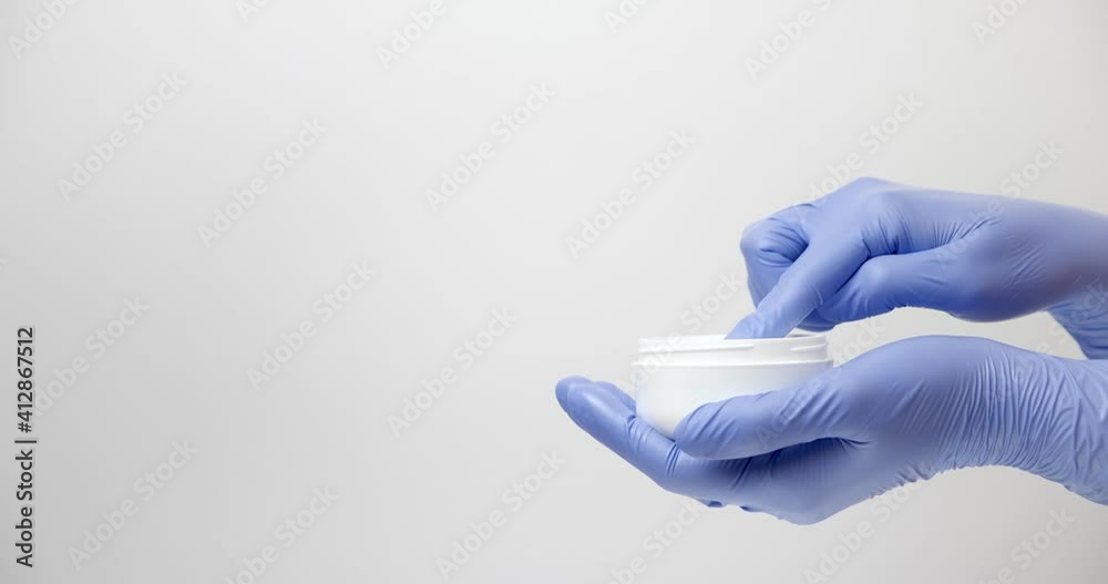 A woman's hand in a medical glove shows a thick hand cream in a jar, on a light background. A woman's hand in a medical glove shows a thick hand cream in a jar, on a light background.