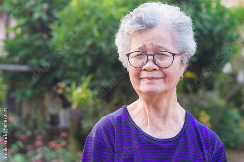 Portrait of a happy senior woman wearing glasses with short gray hair smiling and looking at the camera while standing in a garden. Space for text. Concept of aged people and healthcare