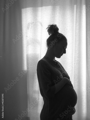 The silhouette of a pregnant girl standing by the window. Black and white photo.