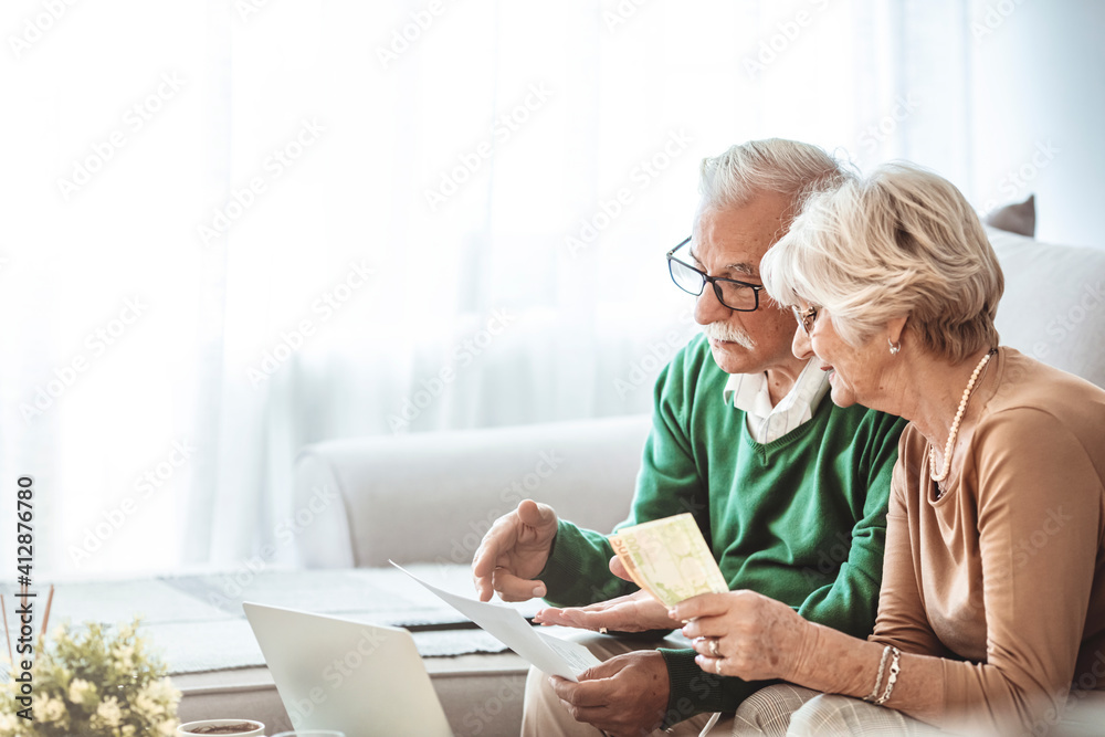Senior couple sitting at a table at home and going through their household finances using a laptop. Senior couple going through bills while using laptop at home. Senior couple sitting at a table at home and going through their household finances using a laptop. Senior couple going through bills while using laptop at home.