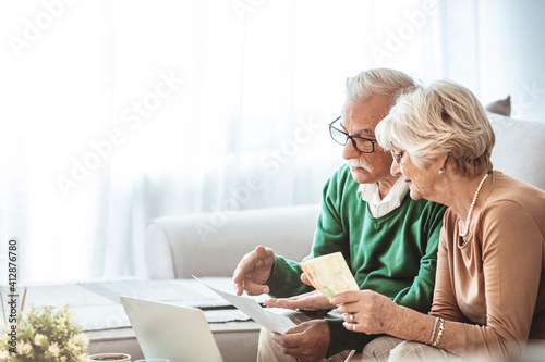 Senior couple sitting at a table at home and going through their household finances using a laptop. Senior couple going through bills while using laptop at home.