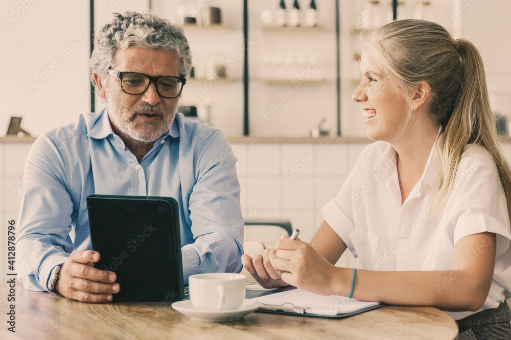 Friendly mature mentor explaining project detail to joyful intern. Business man using tablet and talking to young female colleague. Medium shot. Communication or teamwork concept Friendly mature mentor explaining project detail to joyful intern. Business man using tablet and talking to young female colleague. Medium shot. Communication or teamwork concept
