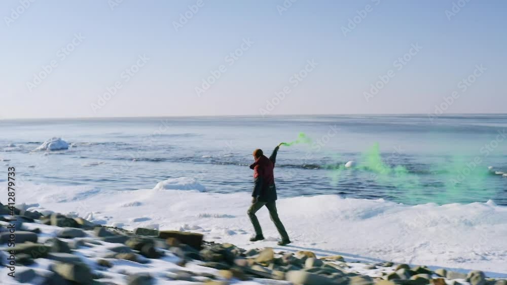 Slow motion aerial view of a man running along the snowy winter beach with green burning flare. Cold blue sea is on the background Slow motion aerial view of a man running along the snowy winter beach with green burning flare. Cold blue sea is on the background