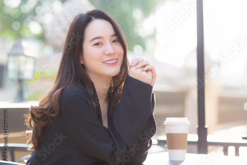 A beautiful Asian woman in a dark blue suit is sitting on a chair and smiling happily. In front of the coffee shop on a sunny morning