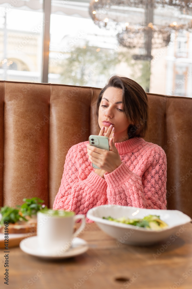 A woman in a restaurant in a warm sweater is looking at something on the phone, a delicious healthy breakfast and matcha latte on the table A woman in a restaurant in a warm sweater is looking at something on the phone, a delicious healthy breakfast and matcha latte on the table