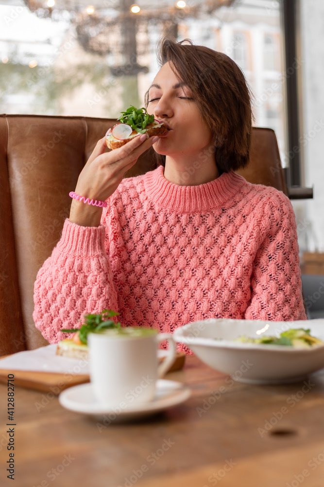 Woman in a restaurant in a cozy warm sweater eating healthy breakfast toast with arugula and salmon Woman in a restaurant in a cozy warm sweater eating healthy breakfast toast with arugula and salmon