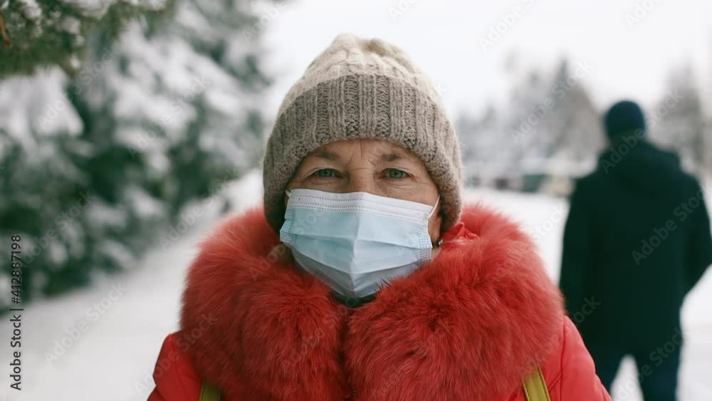 Adult senior woman in protective medical mask looking at the camera in the winter park. Social distancing during Coronavirus Covid 19 pandemic. Beautiful spruce trees branches sprinkled with white Adult senior woman in protective medical mask looking at the camera in the winter park. Social distancing during Coronavirus Covid 19 pandemic. Beautiful spruce trees branches sprinkled with white