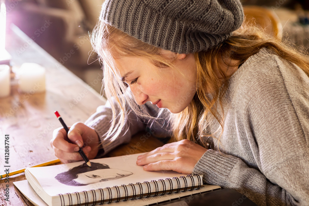 © Mint Images - Teenage girl in a woolly hat drawing with a pencil on a sketchpad.