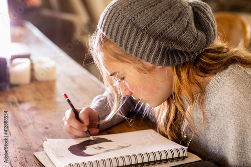 Teenage girl in a woolly hat drawing with a pencil on a sketchpad. photo