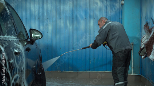 A Caucasian man works in a car wash service. A man in a worker's uniform washes the car thoroughly by spraying it with a water pistol. photo